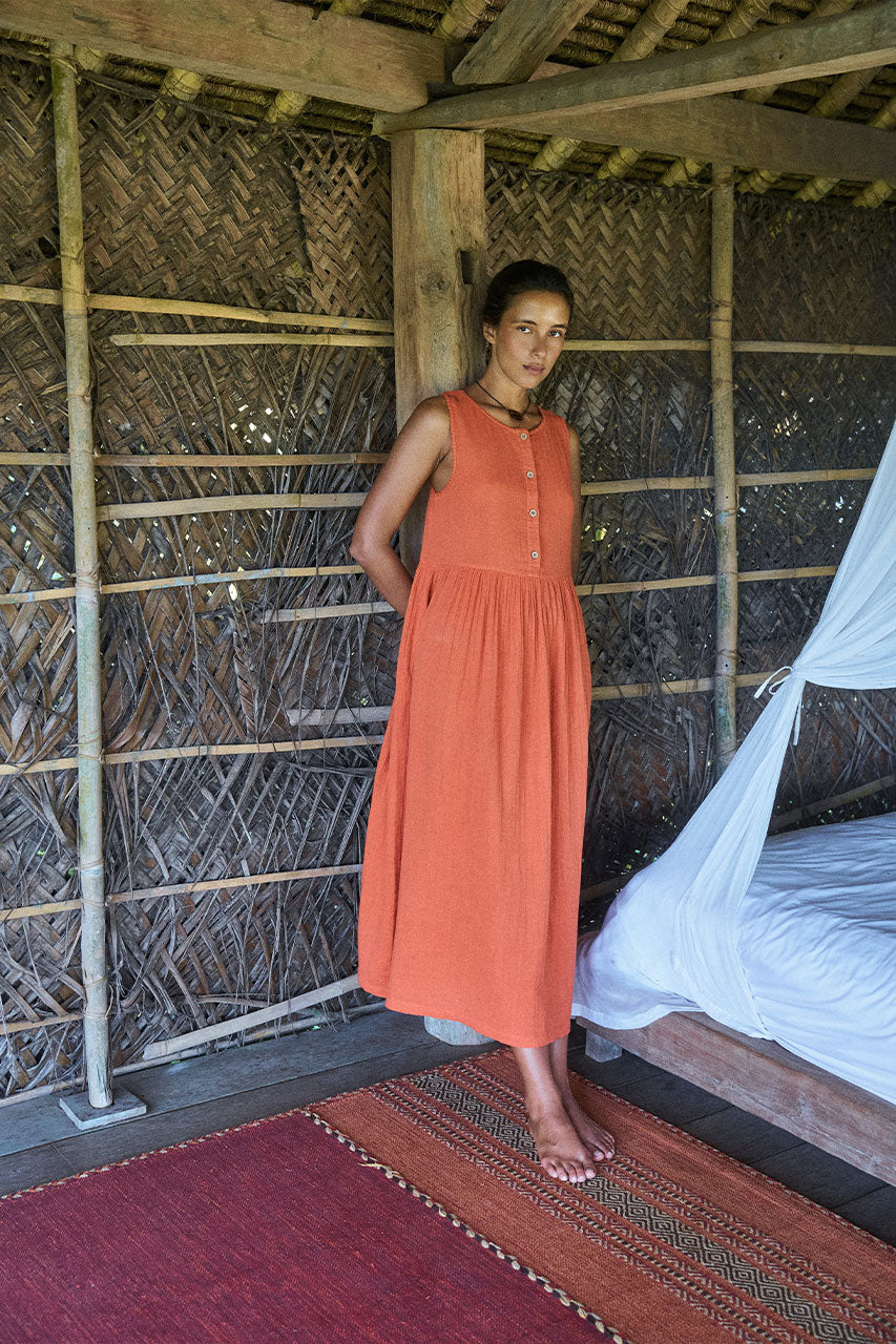 Nadi Dress Terracotta — full length front view standing in rustic bamboo hut beside bed, terracotta double gauze cotton midi dress with wooden button front and gathered waist | Daughters of India